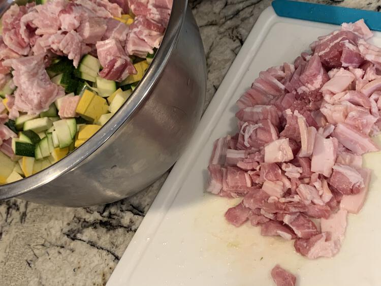 Image shows a variety of chopped squash (both green and yellow skinned) along with raw diced bacon on top. Beside the bowl sits a plastic cutting board containing even more chopped bacon. 