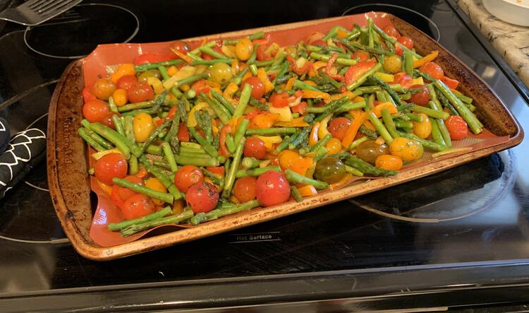 Image shows a variety of cherry tomatoes and pieces of asparagus laid out on a single cookie sheet laid out horizontally on the stove top. Beside it sits and oven mitt and, to the back, a metal flipper to later serve it with. 