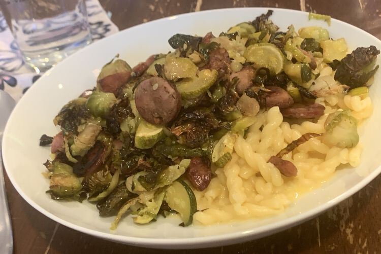 Image shows a large open bowl filled with cheesy pasta and topped with roasted Brussel sprouts, zucchini, and chicken apple sausages. Behind the bowl sits a fabric napkin and a glass of water.  