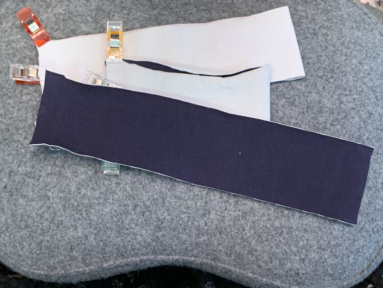 Image shows three bands on the chair. The lower two, a neckband and sleeve cuff, are folded in half lengthwise and clipped on the raw short edges to keep the two layers together. Above them is the other sleeve cuff open and not folded yet. 