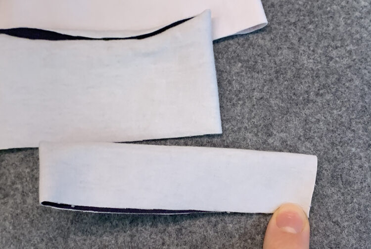 Image shows that cuff from before folded twice creating a four layered rectangle. My finger rests on the corner where both raw edges meet while the folds sit on the top and right side. Beyond it sits the other two bands waiting to be folded a second time. 