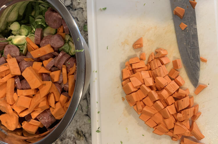 Image shows a white plastic cutting board holding diced yams and a dirtied knife. Beside it sits a large metal bowl partway filled with sliced Brussel sprouts, sliced sausages, and the rest of the yams. 