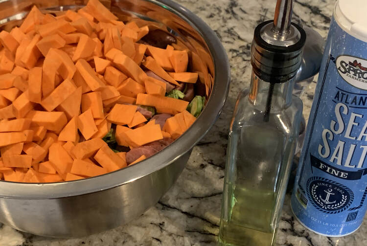 Image shows the same metal bowl although you can hardly see the Brussel sprouts and sausages through the yams. Beside it sits an oil dispenser, sea salt, and a pepper mill. 