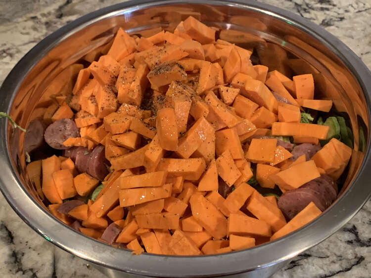 Image shows a bowl of yams with green Brussel sprouts and brown sausages poking through. There is oil and black flecks of pepper in the center back. 