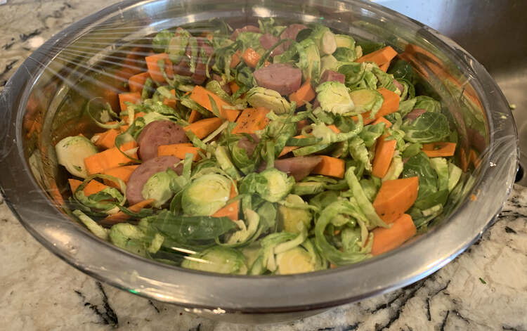 Image shows a metal bowl filled with the evenly mixed Brussel sprouts, yams, sausages, salt, pepper, and avocado oil. Over the top a piece of plastic wrap is stretched across sealing it all in. 