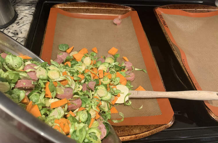 Image shows two cookie sheets lined with an orange bordered Silpat® liner. The metal bowl is angled over the left one as the veggie mix is poured out. A spatula is laid on the cookie sheet waiting to be used to get the last out. 