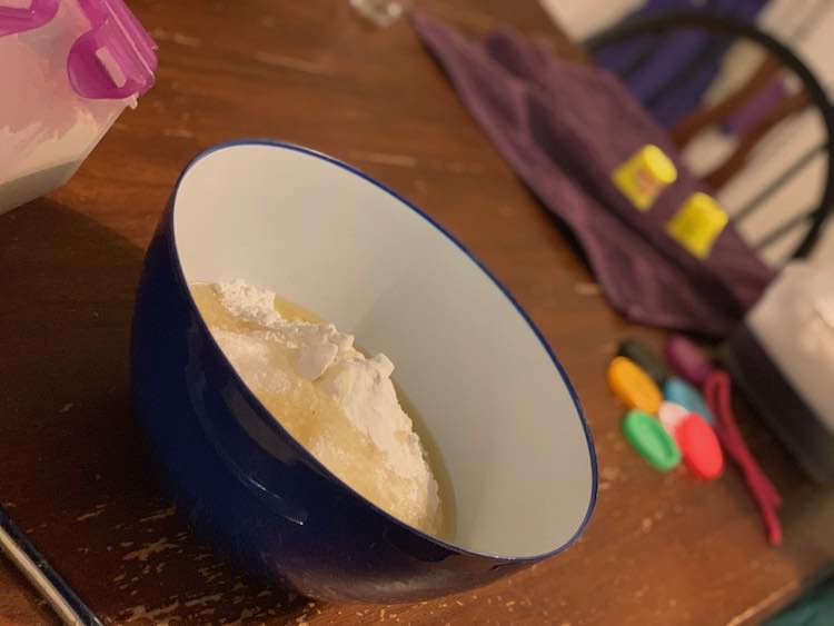 Image shows a blue bowl with a white inside. It's filled with white powder and off white liquid (flour, canola oil, cream of tartar). Off to the side is the purple lidded flour container. In the back is a bin of water, two Play-Doh containers drying on a purple towel, and several colorful lids and a pink pipe cleaner waiting to be cleaned. 