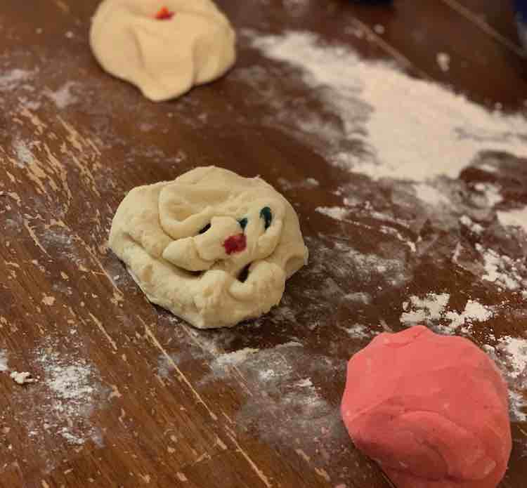 Image shows three different balls of playdough in different states on a flour, white powder, coated table. The front one is fully one shade of pink, the center one is still white but slightly misshapen with drops of blue and red on it (for purple), the back ball is more nest shape and blurred with yellow dye in the center. 