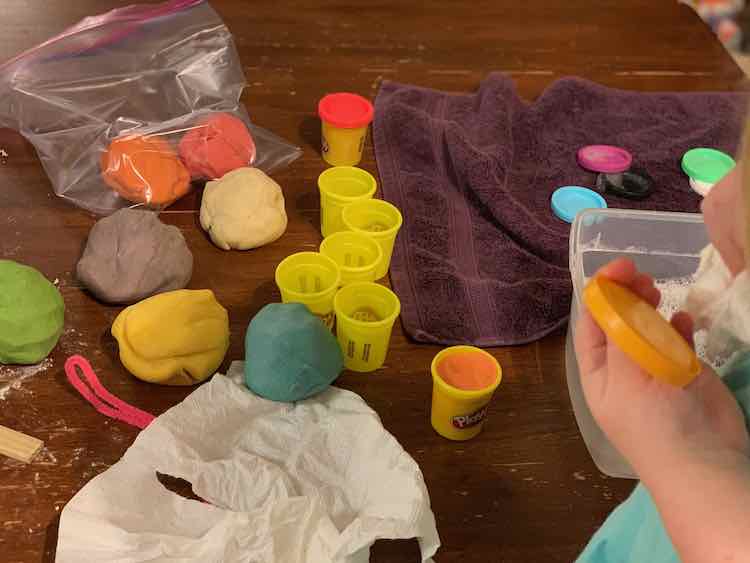 Image shows Ada washing a yellow Play-Doh lid with a white washcloth as a bin of soapy water and some soapy lids on a purple towel sit behind her. Beside that is a lidded red Play-Doh container, several empty and dried containers, and one container filled with orange playdough. Beside this sits several large balls of playdough and behind that a freezer bag with a smaller ball of pink and orange lay the remnants of the containers. 