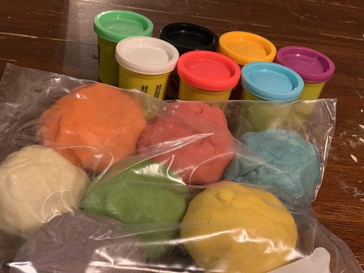 Image shows a clear freezer bag filled with seven different balls of playdough. Behind it sits the seven containers of Play-Doh filled with fresh playdough. 