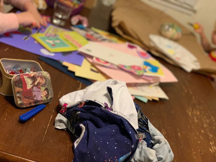 Image shows the dress bunched up on the table with sewing clips attached and black thread sewing. The seam ripper and container of sewing clips sits beside it. In the back and slightly blurred you can see one kid painting a white pumpkin and another cutting up leftover greeting cards. 