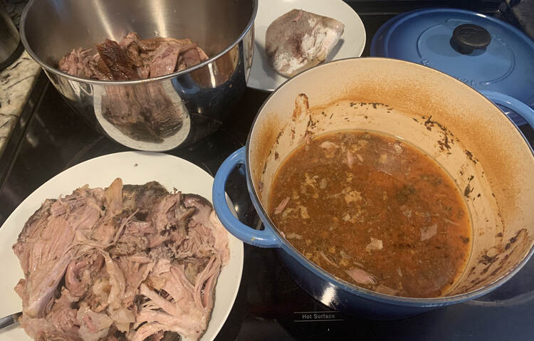 Image shows a white wide bowl and metal stand mixer bowl both with chunks of meat in it. To the right is a dutch oven with a skim of meat juice at the bottom, the lid off to the side, and a saucer with the bone on it. 