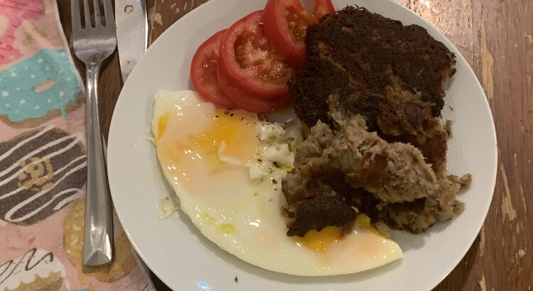 Image shows a white plate topped with brown and black hash, red sliced tomatoes, and fried eggs. Off to the side is a cloth napkin, fork, and knife. 