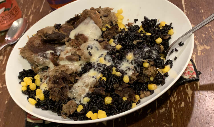 Image shows broken up pork hash, purple rice, and yellow corn all mixed up in a bowl with melted white cheese overtop. There's a spoon sticking out of the mixture. 