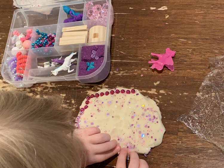 Image shows a glittery circle of white playdough with pink mardi gras style beads forming a fence around the outside. The rest of the kit sits open in the background. 