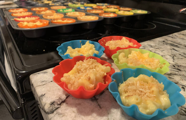 Image shows five mac and cheese muffins sitting on the counter while the rest of the muffins tins sit cooling in the background.