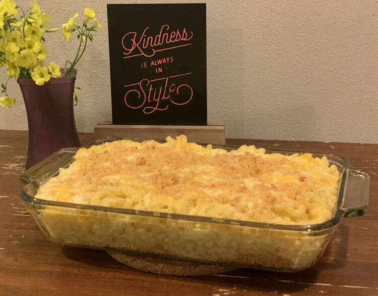 Image shows the mac and cheese casserole in a glass pan on a cork trivet. Behind it sits a sign saying "Kindness is always in style" and a purple vase with matching yellow flowers.