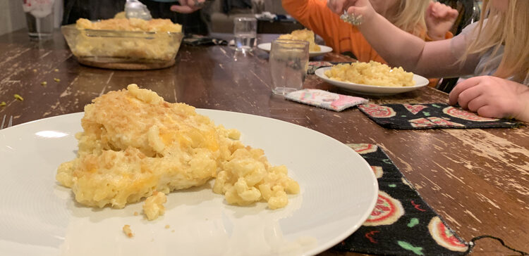 Image shows a serving of mac and cheese on a white plate. In the background you can see three other people sitting around the table eating theirs and the casserole in the center. 