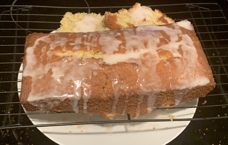 Image shows a loaf, with missing bottom placed beside it, on a black wire cooling rack. White icing is pooled and dripping down the edges as a white plate, below, catches the dribbles. 