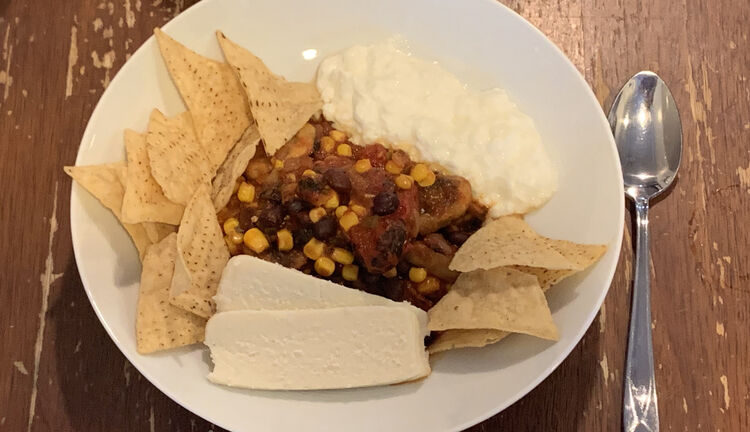 Image shows a white bowl on the table with a metal spoon next to it. The corn bedazzled chili sits in the center of the plate surrounded by yogurt, two piles of tortilla chips, and two slices of cheese. 
