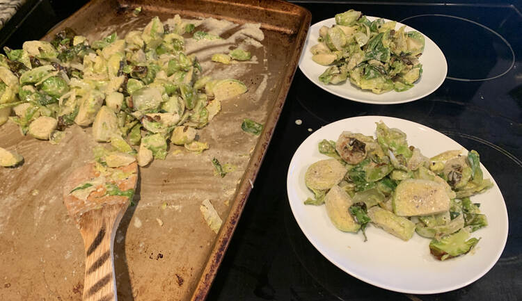 Image shows a cookie sheet with a small pile of white sauce coated Brussel sprouts in a pile and a wooden spatula. Off to the right sits two other plates already dished with the Brussel sprouts. 