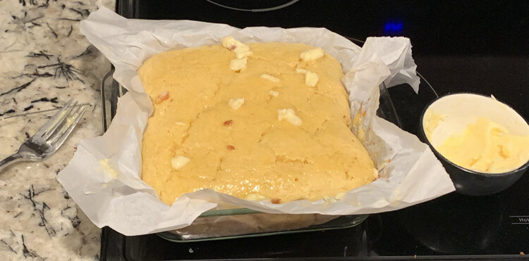 Image shows a square of cornbread with chunks of butter melting overtop. It sits surrounded by parchment paper in a square glass casserole dish sitting beside a black bowl with more sweet butter inside. 