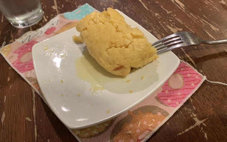 Image shows a corner piece of the cornbread sitting on a square white saucer in a pool of melted butter. A fork lays over the side and a pink donut napkin sits underneath it. A water glass sits in the background.