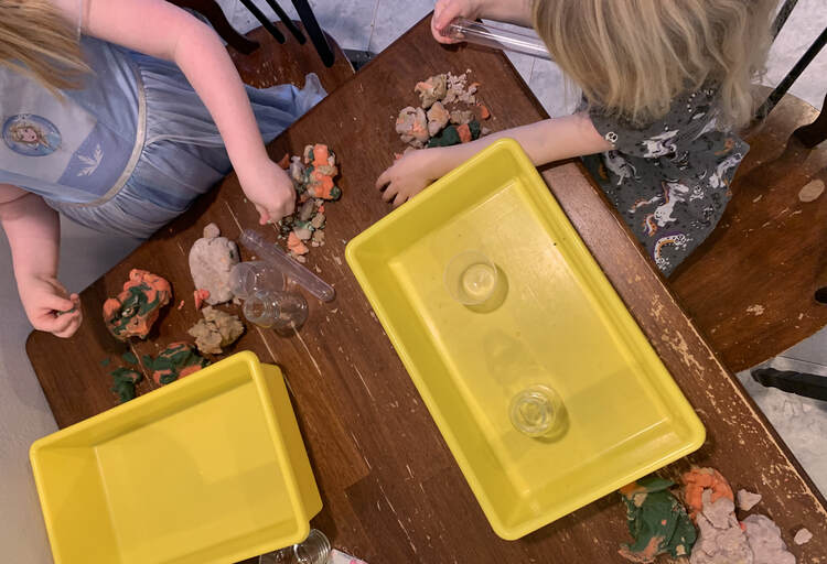 Image shows the kids set up at the end of the table. Both have a yellow bin in front of the and are planning their volcano. 
