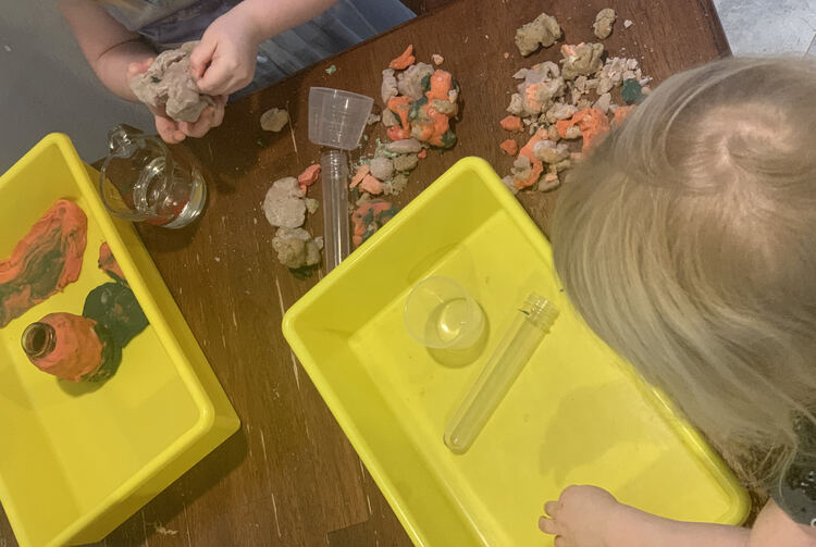 Image shows the girls. Zoey's yellow bin looks empty as her body blocks her glass jar. Ada has started out a playdough floor in the corner of her bin and started encircling her jar with black and pink playdough. She holds some dried grey-ish colored playdough.