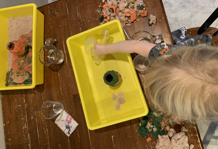 Image shows two yellow bins. The one to the left shows a grey, orange, and black playdough created floor with an orange volcano in the center. Zoey is leaning over the bin to the right moving the plastic containers as her playdough encased volcano sits in a pool of water. 