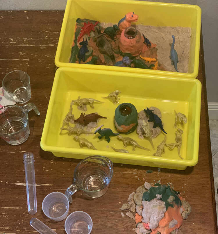 Image shows a view from above both plastic bins holding a dinosaur world. The top bin, further back, is Ada's filled with the playdough floor. There are colorful plastic dinosaurs in the land mostly on the black and orange section while a single Apatosaurus stands on the grey part. Zoey's looks mainly yellow as the only playdough in her bin is the volcano and several rocks. There are three solid colored dinosaurs and the rest are yellow skeletons. One dinosaur is on the pebbles, two on the rock, and the rest are in the water. 