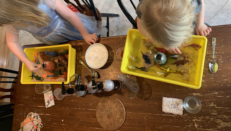 Image is taken from above at both girls' dinosaur worlds side by side. On the left Ada sprinkles a pinch of baking soda into her volcano while Zoey, on the right, plays with a dinosaur first. 