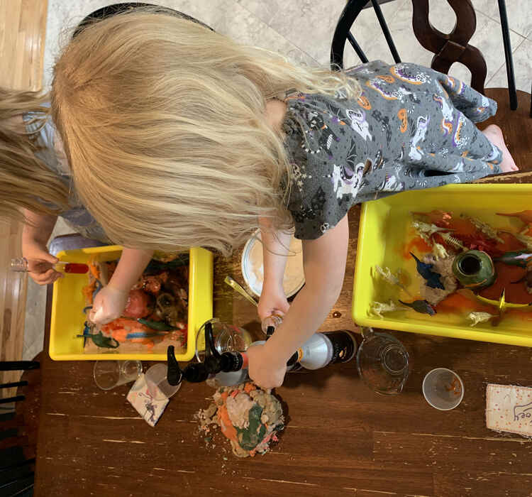 Image shows Zoey standing on her chair pumping yellow dyed vinegar into her plastic vial. Her yellow bin below shows orange lava spreading through the clear water. In the back ground Ada is pouring baking soda into her volcano with one hand and holding a vial filled with red magma in the other.  
