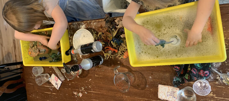 Image shows knocked over vinegar pump bottles, Ada playing in colored vinegar coated playdough dinosaur world, and Zoey with soapy water and cleaner dinosaurs.  