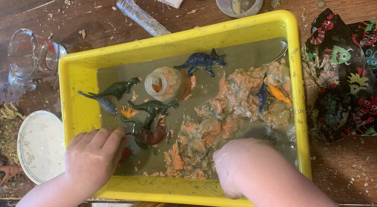 Image shows a closeup of Ada's green black colored liquid covering the orange and black playdough with some dinosaurs, a glass bottle, and a spoon hidden within. 