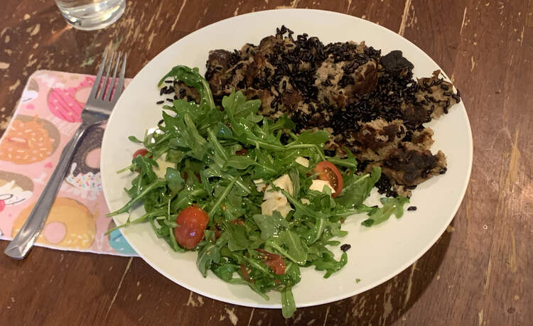 Image shows a white plate with a pink donut napkin beside it and a fork. Cut off on the top left you can see the bottom of a water glass. The plate is half filled with a green, red, and white salad on the one side and a mix of black and brown stuff on the other.