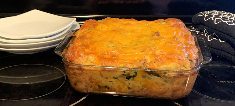 Image shows the same glass casserole dish filled with a peaked dish topped in melted orange cheese. Behind it sits four saucers, to the left, and stacked oven mitts to the right. 