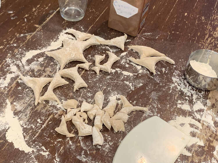 Image shows the table coated in flour and dough remnants. Off to the side sits a measuring cup with more flour, a mix of cinnamon and sugar, a dirty glass, and a plastic bench scraper. 