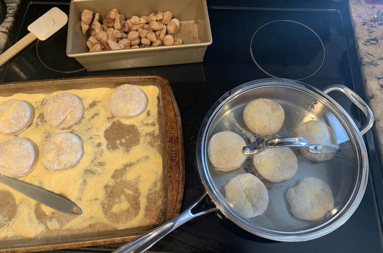 Image shows the top of the stove. To the left sits a cornmeal coated cookie sheet half filled with raw dough circles. To the right sits six English muffins frying in oil with a lid overtop. In the back sits the baked sugar and cinnamon sprinkled dough bits. 