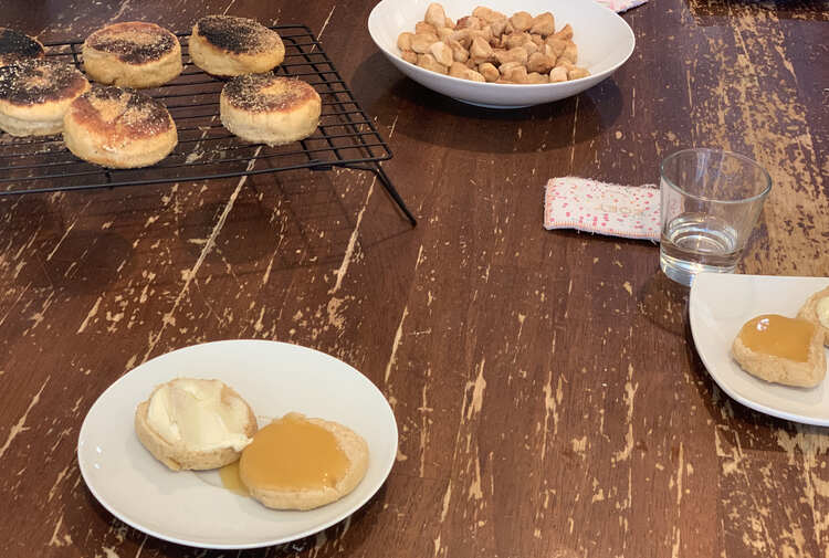 Image shows two saucers with a sliced open English muffin topped with cream cheese, on one, and lemon curd on the other half. In the background sits some burnt English muffins cooling on a wire rack and the bowl of baked scraps. 