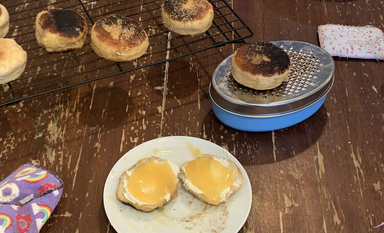 Image shows a sliced English muffin on a saucer topped with cream cheese and lemon curd. In the background sits a English muffin resting on a zester with a wire rack holding the rest. 