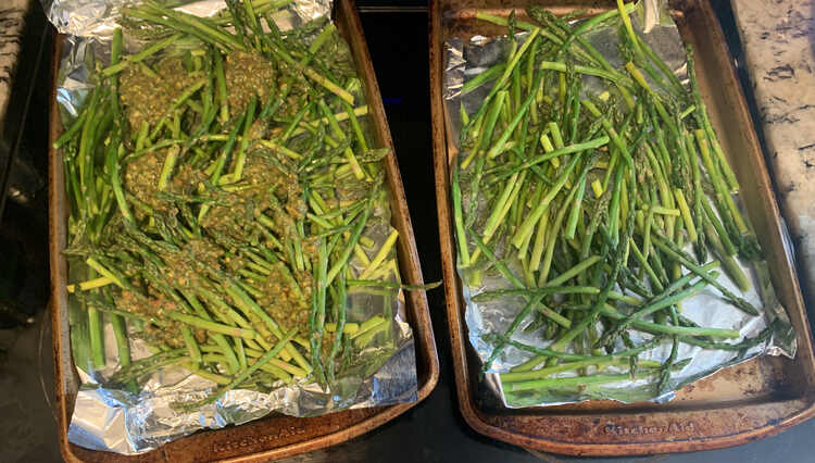Two tinfoil lined cookie sheets lay out on the stove with bright green asparagus on it. The one on the left has dollops of a different hued green all over it. 