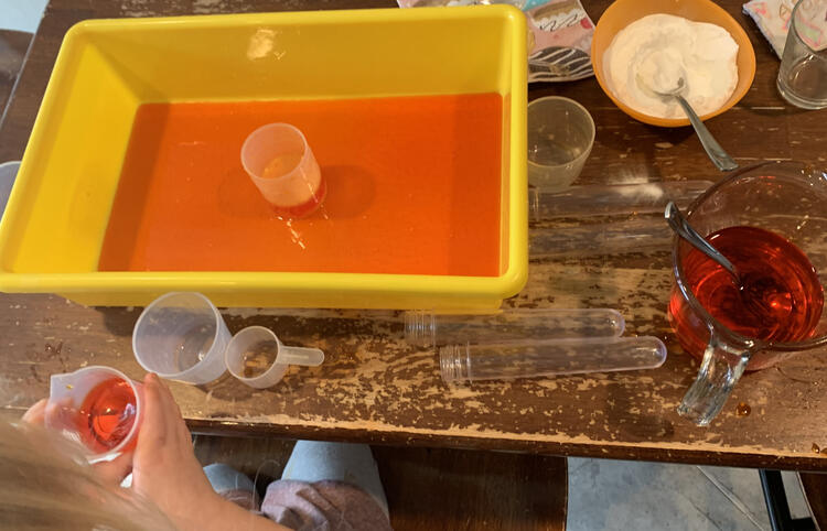 Image is taken from above looking down at the table. On it is a yellow rectangular bin with a layer of orange liquid at the bottom and a plastic measuring cup in the center. Around it sits an orange bowl with baking soda and a spoon, several more plastic cups and vials, and a glass measuring jug half filled with orange vinegar and a spoon. Zoey's hands are showing at the bottom of the photo grasping a plastic beaker of orange liquid.