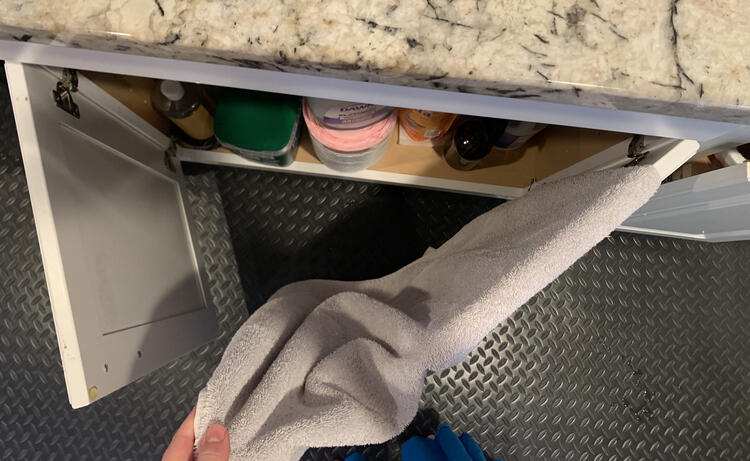 Image is taken from above looking down at the kitchen counter and showing the opened cupboard doors under the sink. The towel is folded over one of the cupboard doors while my hand holds the end. The other door is left alone. 