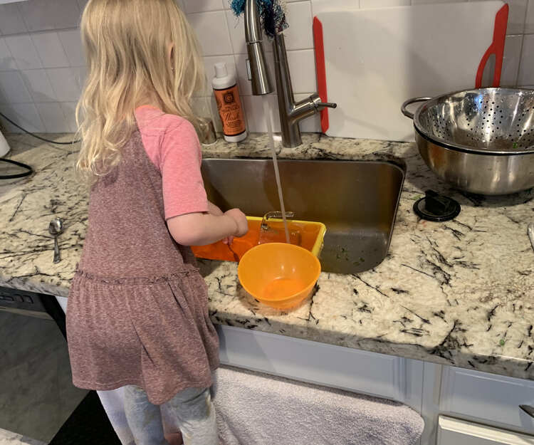 Image shows Zoey standing on chair (chair cut off) in front of the kitchen sink and towel covered cupboard doors. In the sink the yellow bin is full with orange tinted water as more water runs into it from the faucet. 