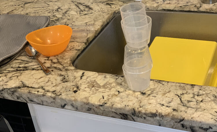 Image shows a stack of several plastic measuring cups, leftover from laundry detergent, stacked in front of the kitchen sink. In the sink sits the upside down yellow bin while to the left sits an orange bowl and metal spoon beside the drying pad. 