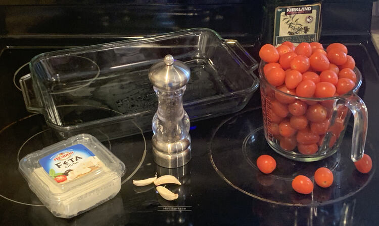 Image shows all the ingredients I'm about to assemble laid out around my glass casserole pan on the stove top. This includes (from top right clockwise) a bottle of olive oil, an overflowing four cup measuring jug of cherry tomatoes, a pepper mill, three garlic cloves, and a plastic container holding a block of feta. 