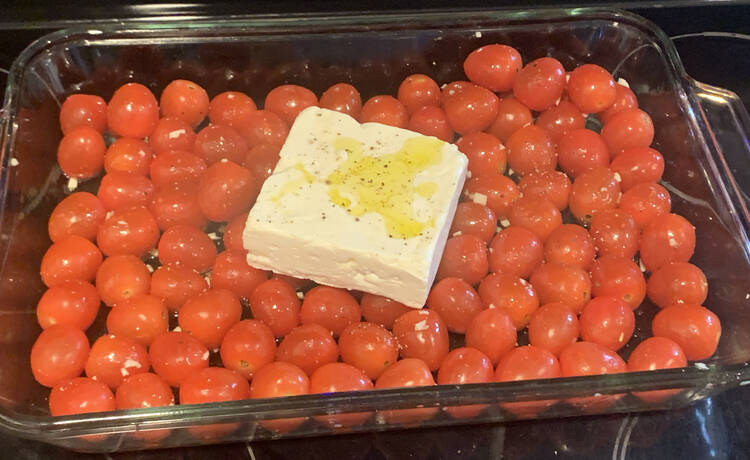 Image shows a glass casserole dish filled with a single layer of oiled cherry tomatoes with a white block of feta cheese in the center. The cheese has a yellow pool of oil and black pepper flecks. Amongst the tomatoes you can see the odd black fleck and minced garlic bits.