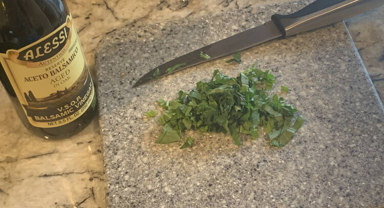 Image shows a cutting board with a pile of slivered green basil next to a used knife. Beside the cutting board sits a bottle of aged balsamic vinegar. 