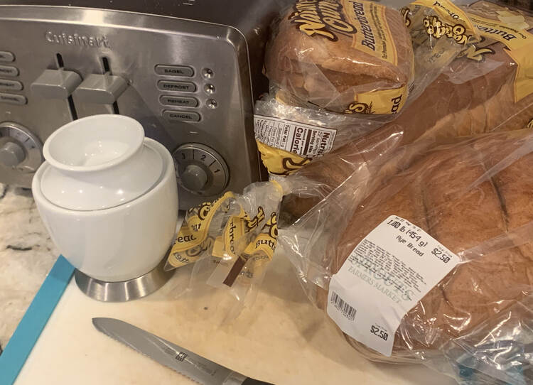 Image shows a silver toaster beside a bag of sliced butterbread. In front of it sits a while unsliced rye bread loaf, a white butter crock, a plastic cutting board, and a serrated bagel knife. 